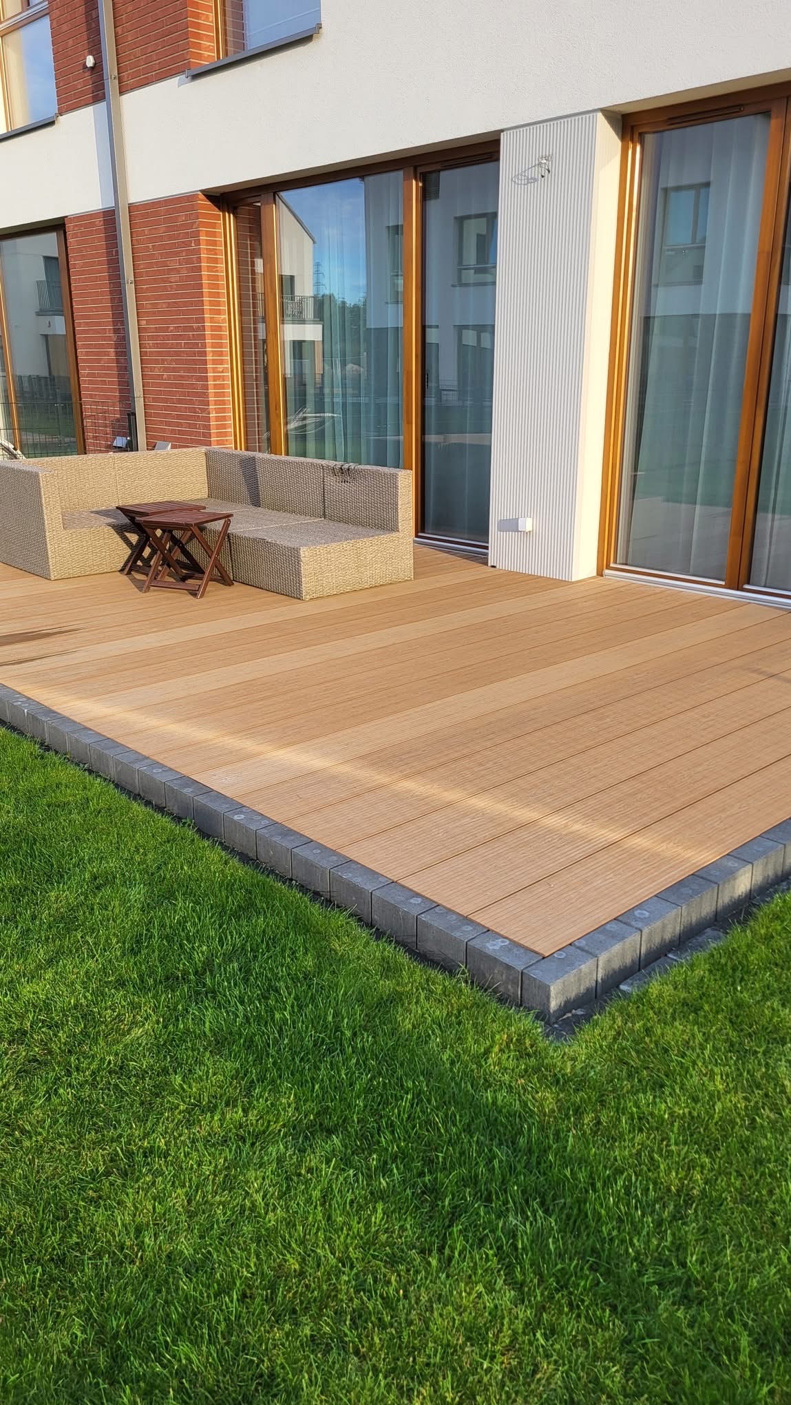 Nowoczesny taras kompozytowy z obrzeżem z kostki, przylegający do budynku z dużymi oknami, z narożną sofą i stolikiem kawowym. Widoczny fragment trawnika.