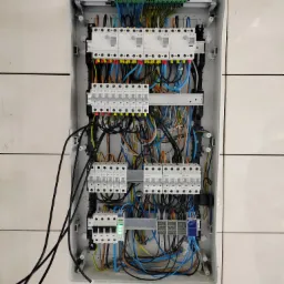 Otwarta skrzynka elektryczna z widocznymi bezpiecznikami, przewodami w różnych kolorach i listwami zaciskowymi, zamontowana na ścianie wyłożonej białymi płytkami. Widoczne cztery czarne przewody...