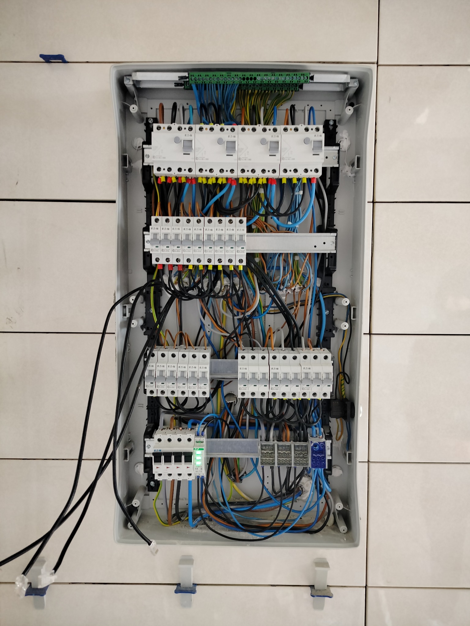 Otwarta skrzynka elektryczna z widocznymi bezpiecznikami, przewodami w różnych kolorach i listwami zaciskowymi, zamontowana na ścianie wyłożonej białymi płytkami. Widoczne cztery czarne przewody...