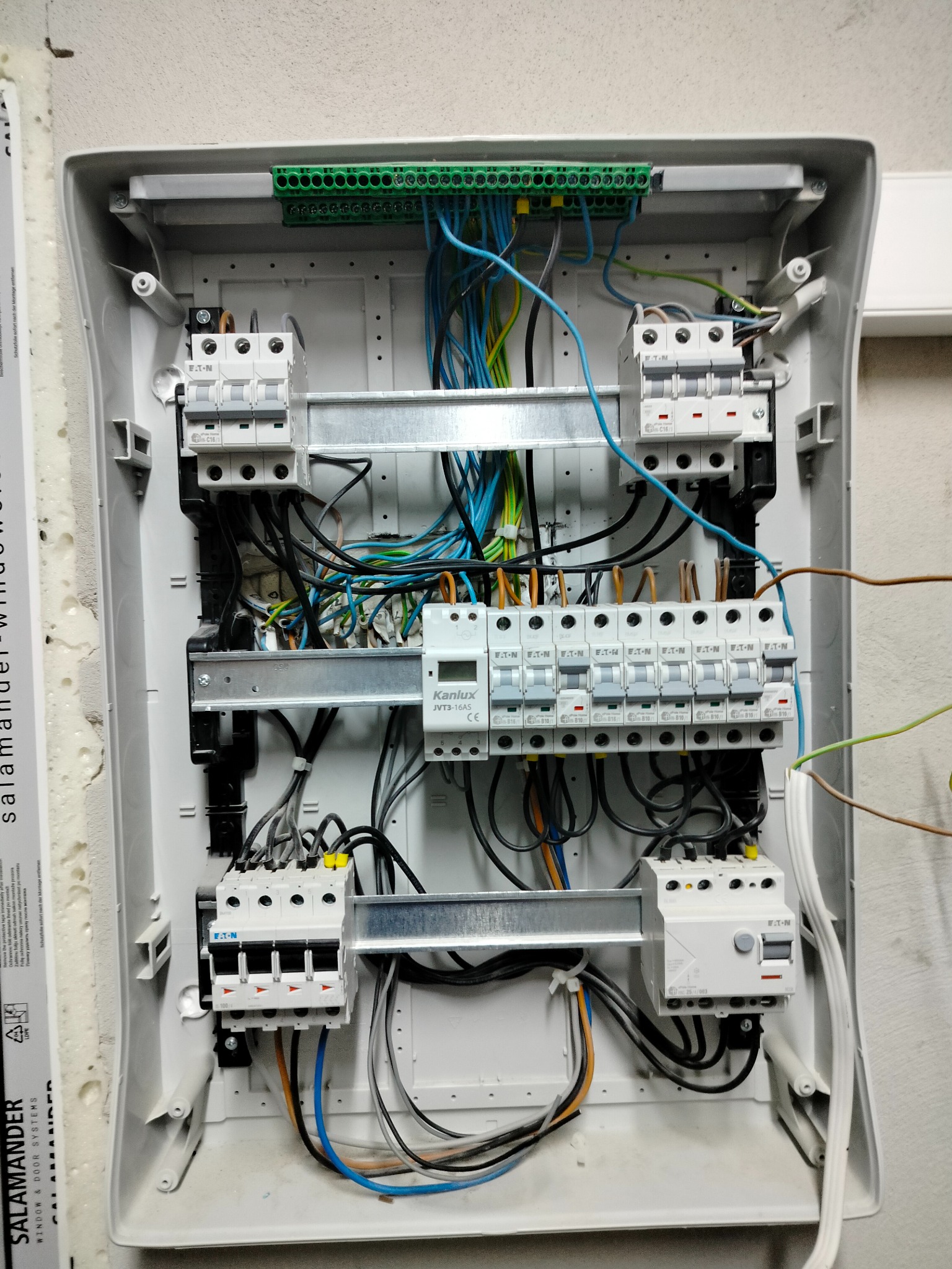 Otwarta skrzynka bezpiecznikowa z widocznymi bezpiecznikami Eaton, listwą zaciskową, przewodami w różnych kolorach i szyną montażową, zamontowana na ścianie obok okna Salamander.