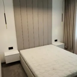 Nowoczesna sypialnia z beżowym panelem tapicerowanym za ł&oacute;żkiem, minimalistycznymi szafkami nocnymi i wiszącymi lampami z czarnymi i złotymi elementami.