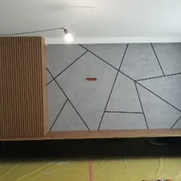 Nowoczesna szafa na wymiar z drewnianymi lamelami i czarnymi frontami, na tle ściany z geometrycznym wzorem, widoczna instalacja elektryczna