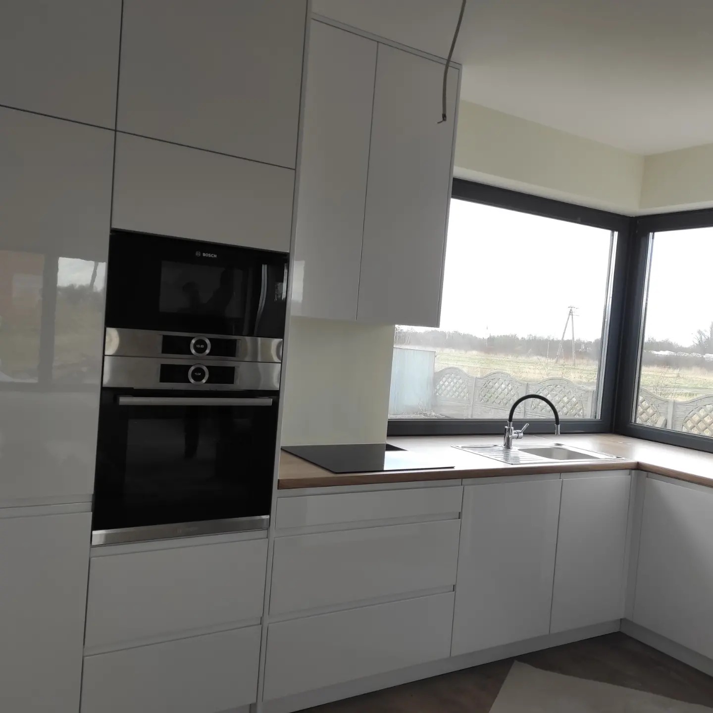 Nowoczesna kuchnia z białymi, błyszczącymi frontami, zabudowanym piekarnikiem i płytą grzewczą, jasnym blatem drewnopodobnym oraz dużym oknem z widokiem na zewnątrz.