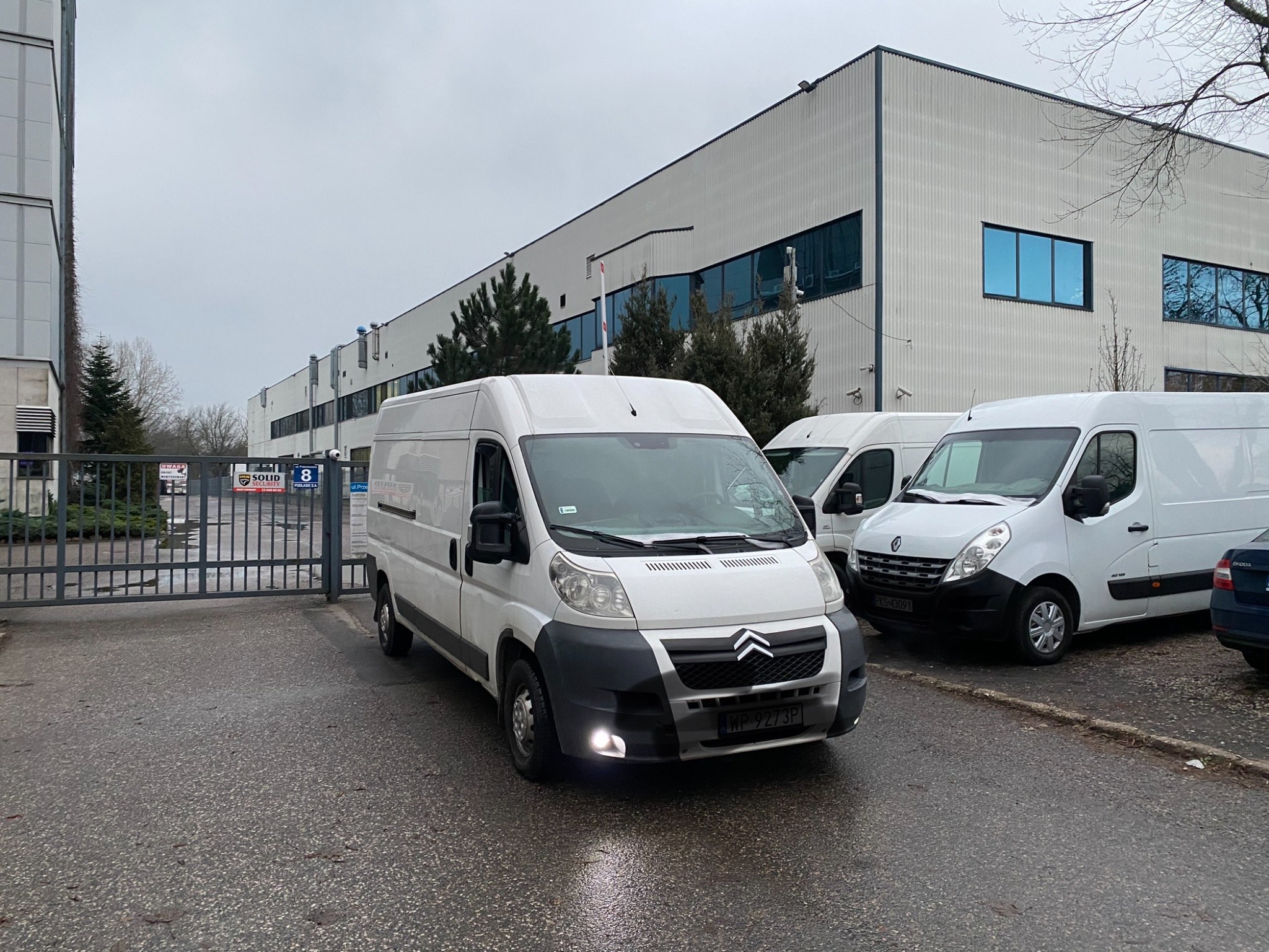 Biały Citroen Jumper z włączonymi światłami mijania, zaparkowany na mokrym asfalcie przed szarą bramą wjazdową i budynkiem magazynowym z niebieskimi oknami w tle. Obok stoją inne białe busy.