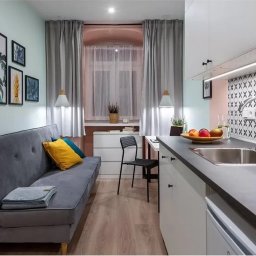A.W. Construction Ariel Włodarczyk - Aranżacja małego mieszkania z aneksem kuchennym: szara sofa z kolorowymi poduszkami, dekoracyjne obrazy na ścianie, białe szafki kuchenne z czarnym blatem i mozaiką na ścianie.