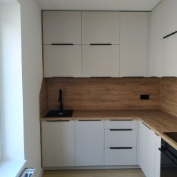 A.W. Construction Ariel Włodarczyk - Nowoczesna kuchnia w bieli i drewnie: minimalistyczne szafki z czarnymi uchwytami, drewniany blat i panel nad blatem, czarny zlew i bateria, jasna podłoga, widok na grzejnik pod oknem.