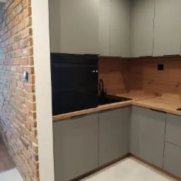 Nowoczesna kuchnia w odcieniach szarości z drewnianym blatem i ceglaną ścianą, czarna armatura i sprzęt AGD, minimalistyczny design, oświetlenie sufitowe.