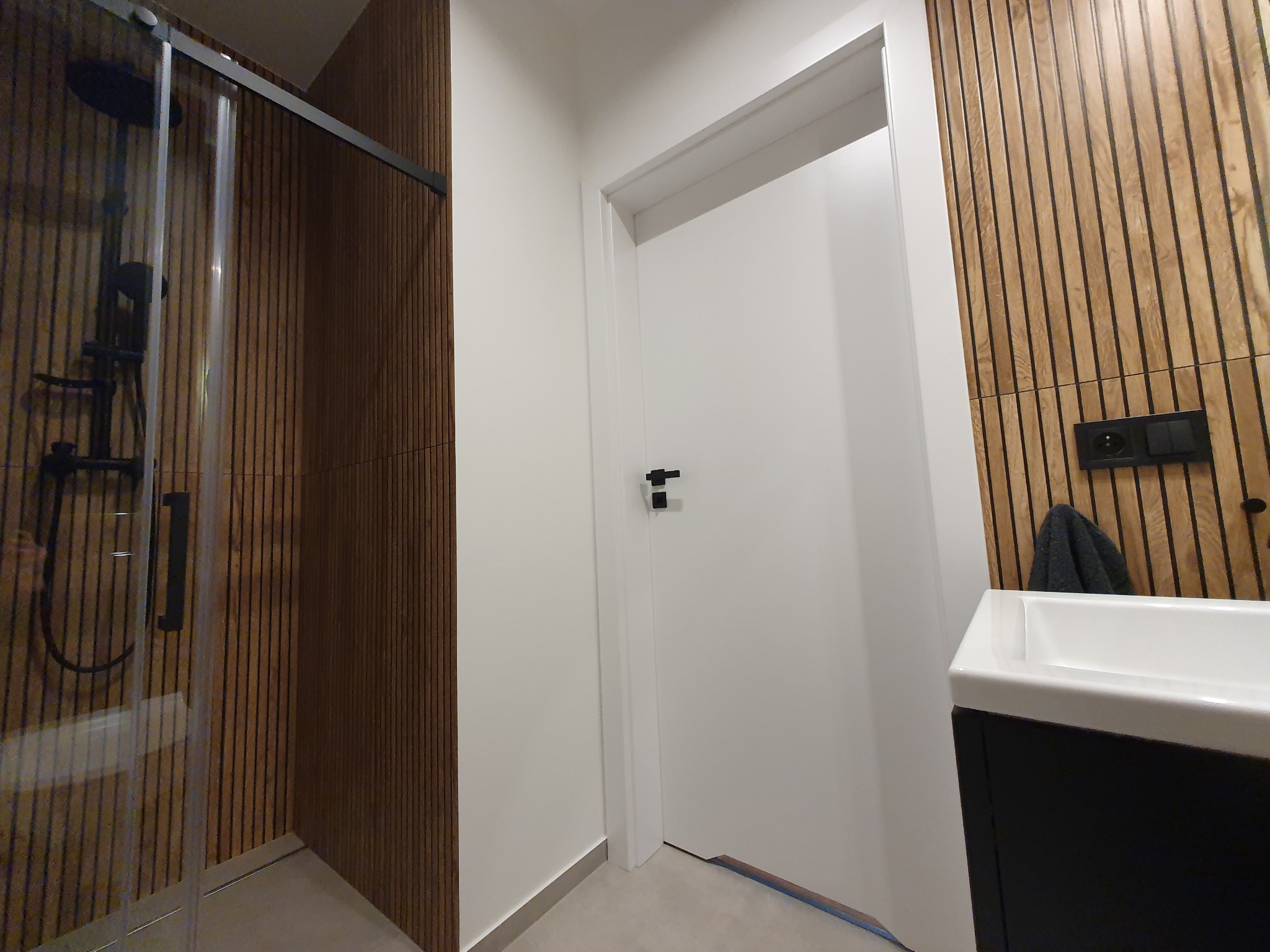 Nowoczesna łazienka z kabiną prysznicową typu walk-in, drewnianymi panelami ściennymi, białą umywalką nablatową i czarnymi detalami, takimi jak bateria i włączniki światła.