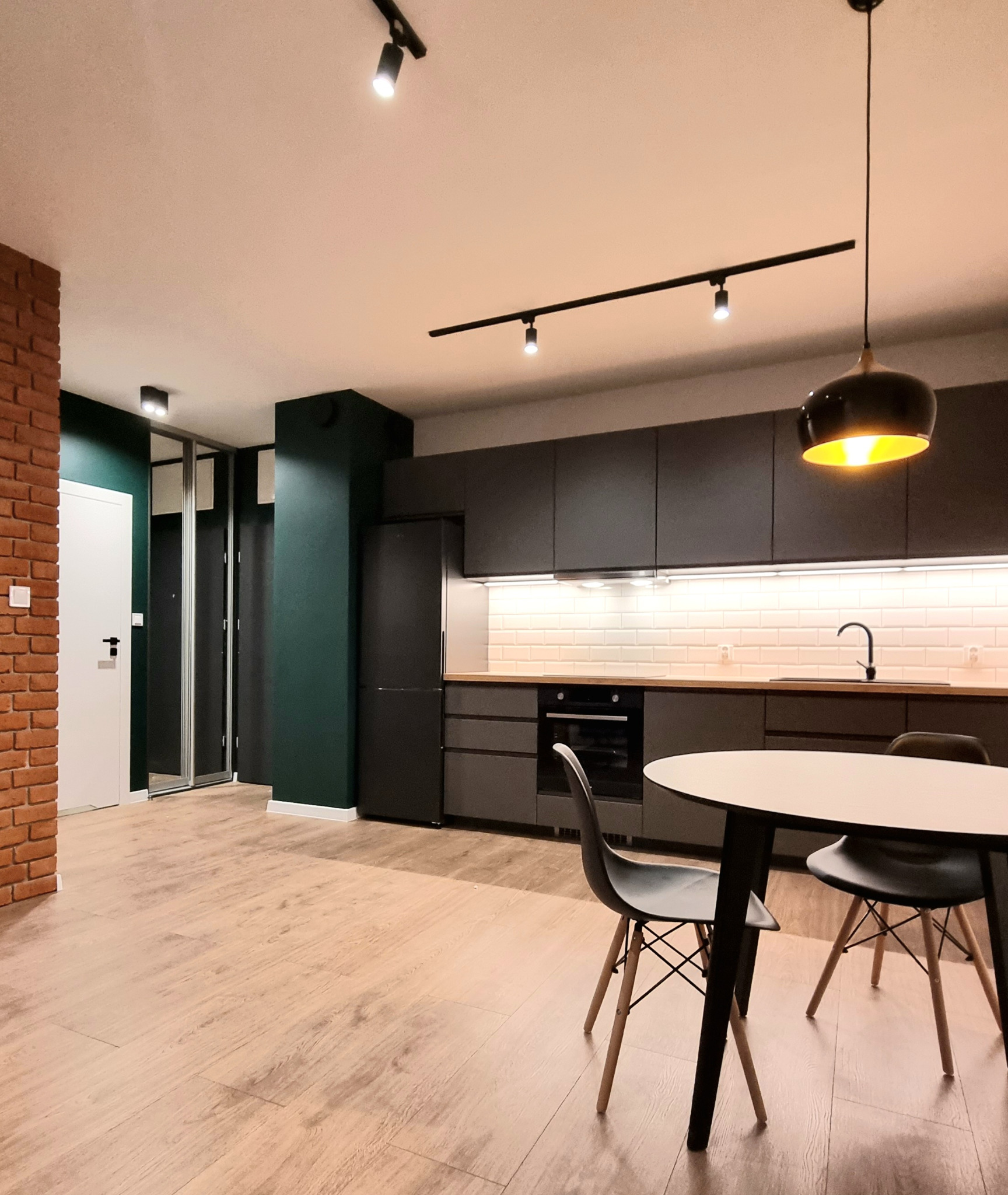 Nowoczesna kuchnia w odcieniach szarości i zieleni z ceglanym akcentem, oświetleniem punktowym i wiszącą lampą nad okrągłym stołem z krzesłami typu Eames.