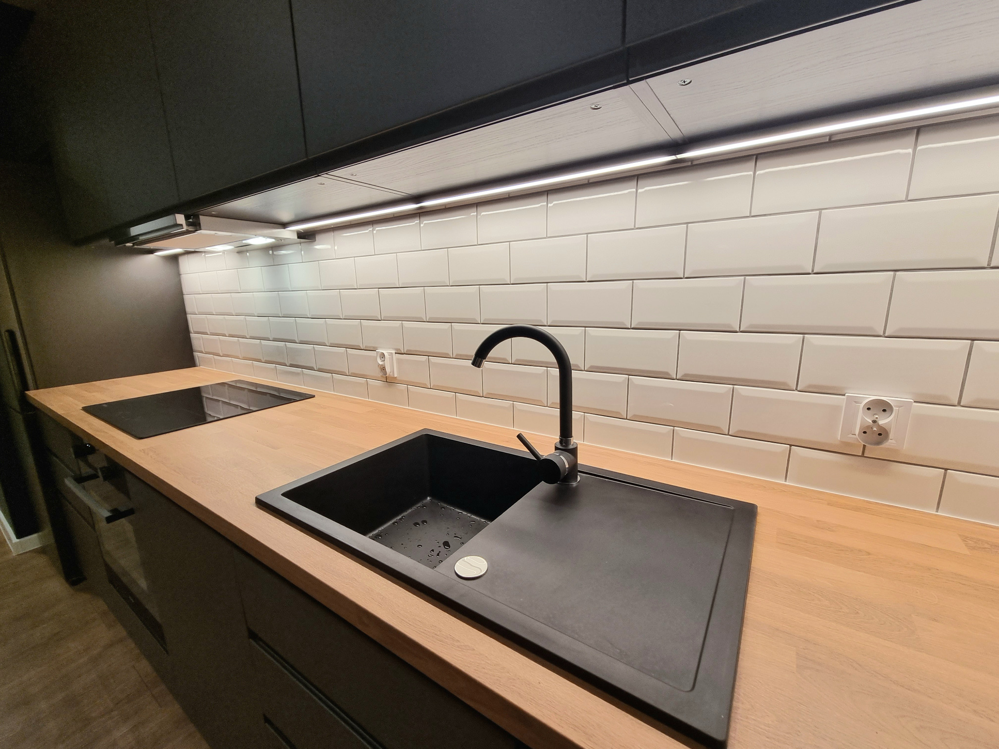 Nowoczesna kuchnia z czarnym zlewozmywakiem, drewnianym blatem i białymi, wypukłymi płytkami typu metro, oświetlona listwą LED pod szafkami.