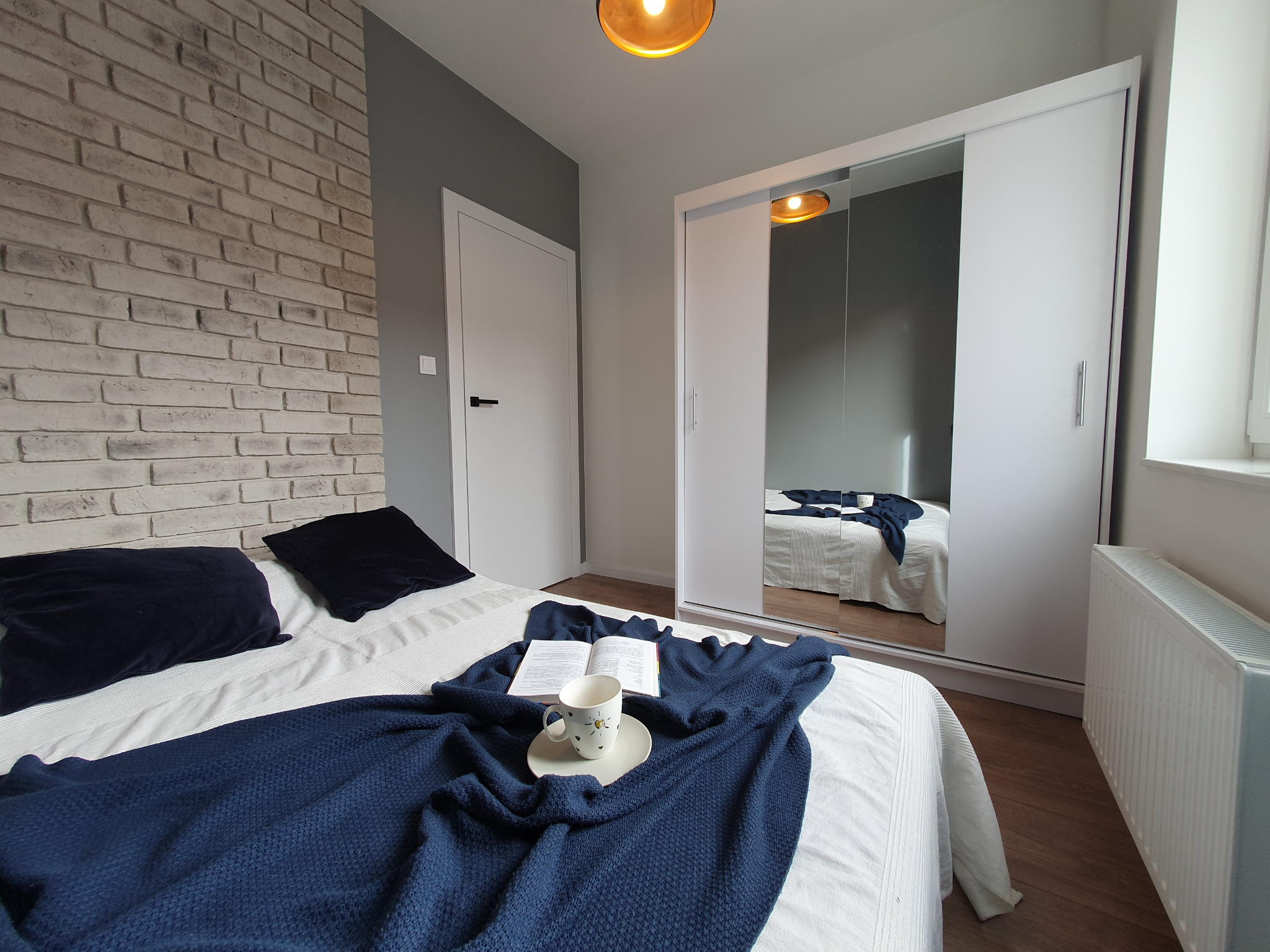 Nowoczesna sypialnia z łóżkiem z ciemnoniebieską narzutą, otwartą książką i kubkiem na talerzyku, ściana z cegły dekoracyjnej, biała szafa przesuwna z lustrami i białe drzwi.