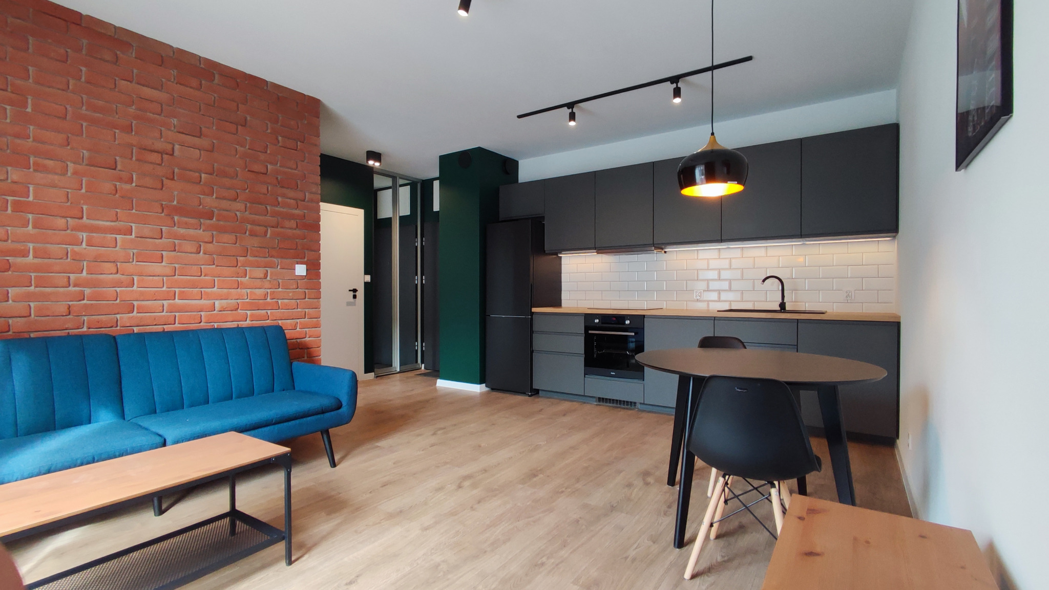 Nowoczesne wnętrze mieszkania z ceglaną ścianą, turkusową sofą i szarą kuchnią z drewnianym blatem oraz okrągłym stołem