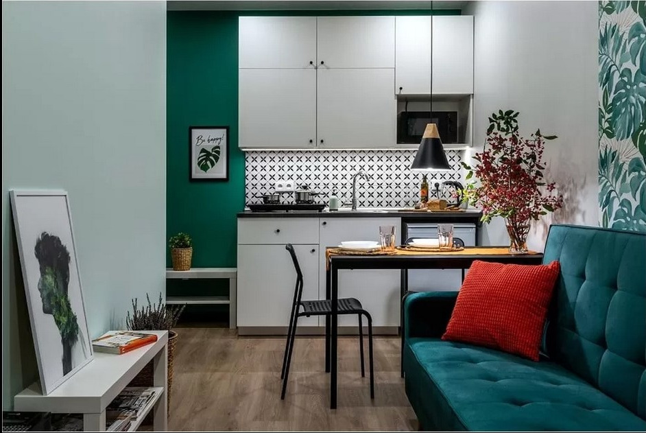 Wnętrze kawalerki z aneksem kuchennym: białe szafki, czarno-biała mozaika nad blatem, turkusowa ściana i sofa z czerwoną poduszką, dekoracje roślinne i grafiki na ścianach.