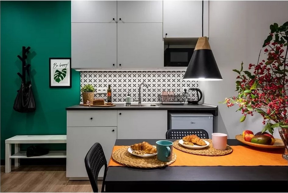 Nowoczesna, urządzona kuchnia w odcieniach szarości i zieleni z czarnymi akcentami, widok na stół z croissantami, dekoracyjną lampę i ścianę z wzorzystymi płytkami.