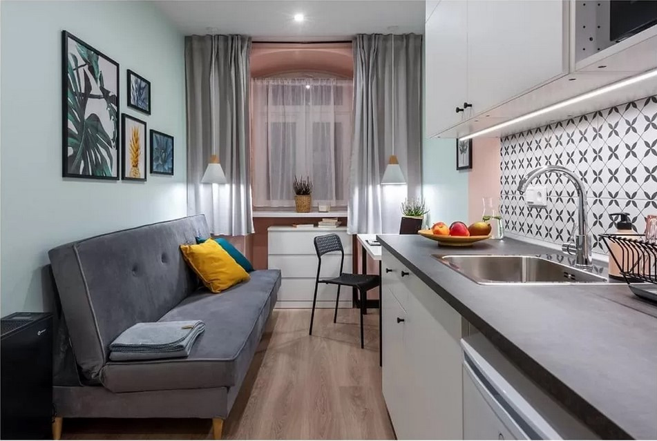 Aranżacja małego mieszkania z aneksem kuchennym: szara sofa z kolorowymi poduszkami, dekoracyjne obrazy na ścianie, białe szafki kuchenne z czarnym blatem i mozaiką na ścianie.