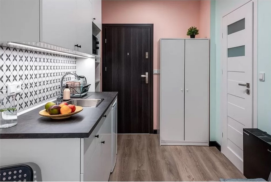 Nowoczesna kuchnia z ciemnym blatem, wzorzystą mozaiką nad zlewem, drewnianą podłogą i kolorowymi ścianami w odcieniach różu i zieleni, z widocznymi owocami w misce.