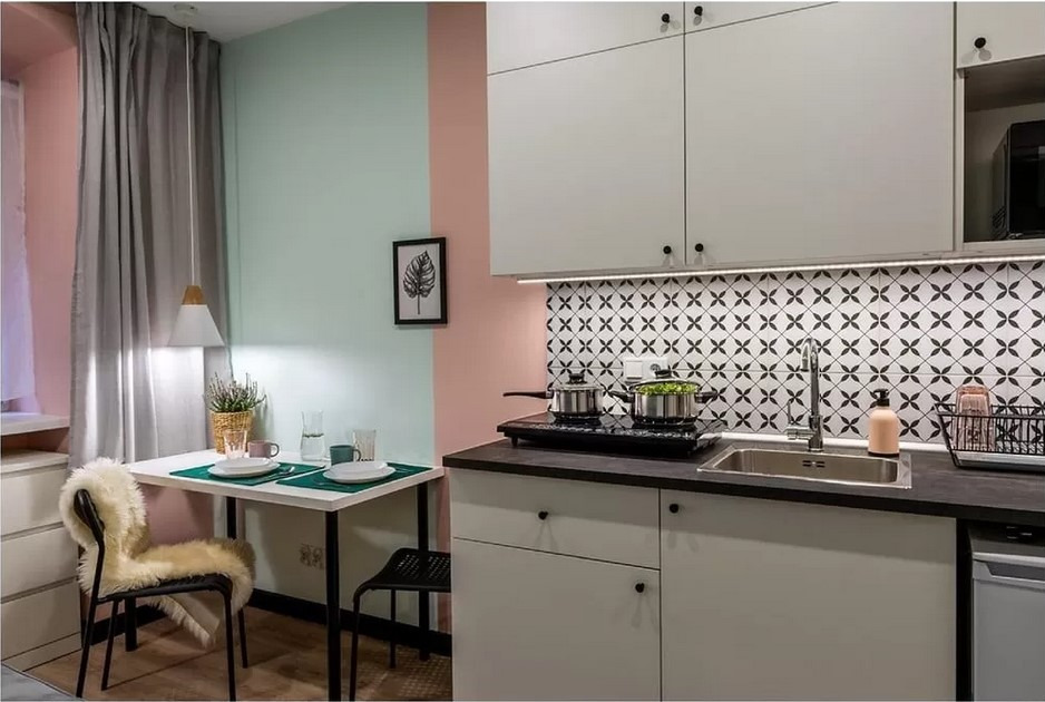Nowoczesna, pastelowa kuchnia z białymi szafkami, wzorzystą mozaiką nad blatem, indukcją, zlewozmywakiem i małym stolikiem jadalnym z futrzanym krzesłem.
