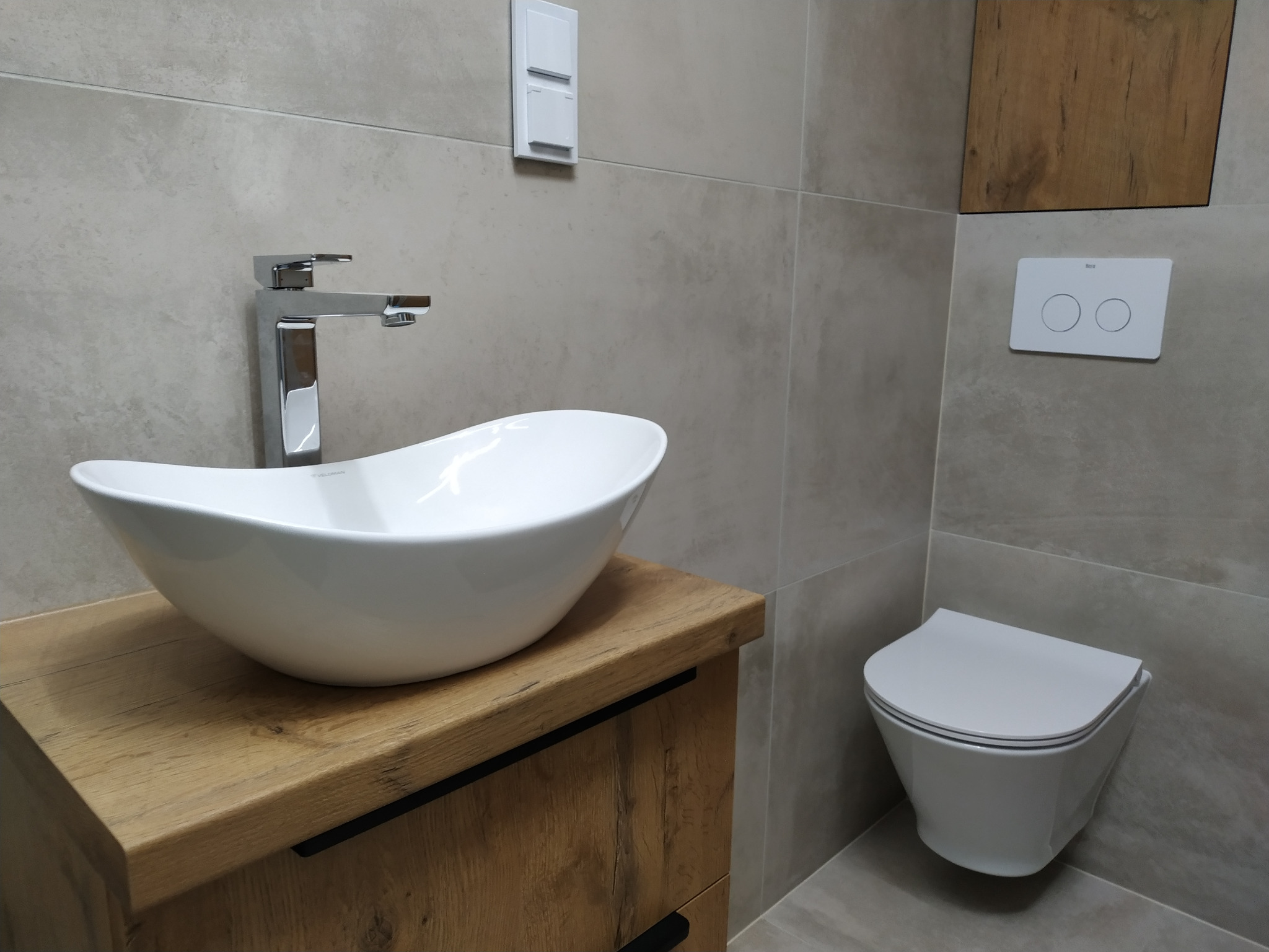 Nowoczesna łazienka w odcieniach szarości z umywalką nablatową w kształcie łzy na drewnianej szafce z czarnymi uchwytami, chromowaną baterią i podwieszaną toaletą z przyciskiem spłukującym Roca.