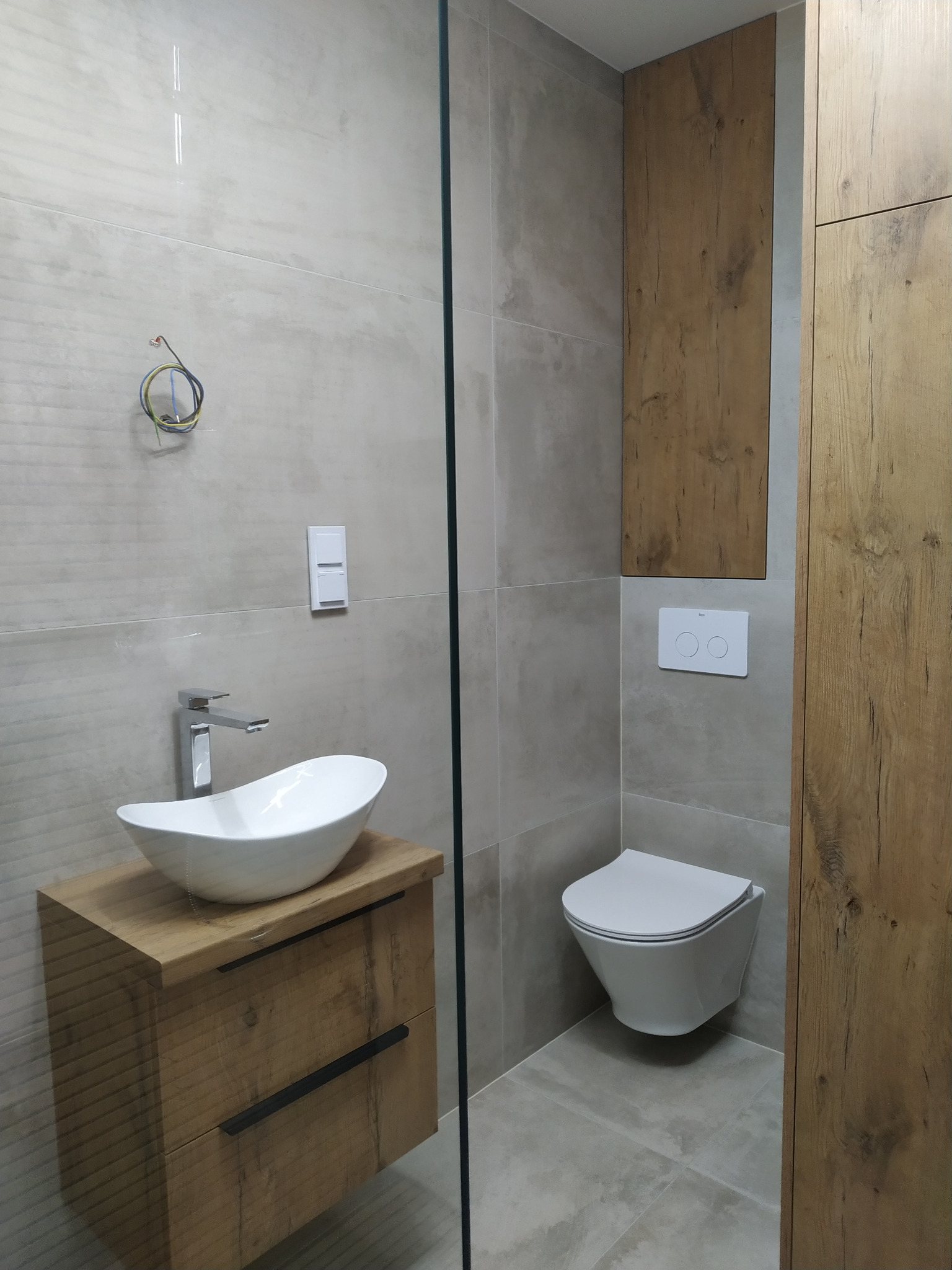 Nowoczesna łazienka w odcieniach szarości i drewna z wiszącą toaletą, umywalką nablatową o owalnym kształcie na szafce z drewnianym frontem i wystającymi przewodami elektrycznymi na ścianie.
