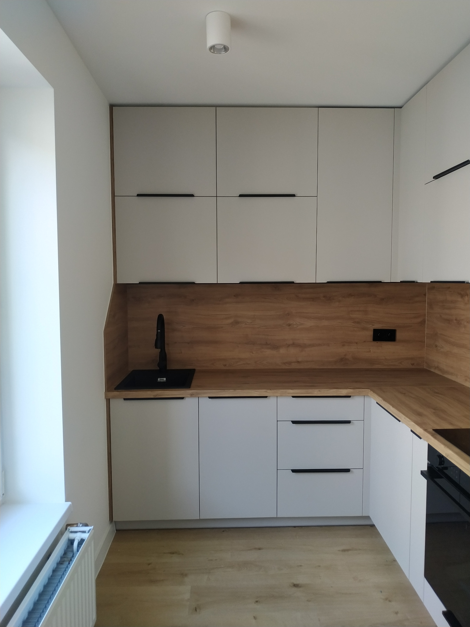 Nowoczesna kuchnia w bieli i drewnie: minimalistyczne szafki z czarnymi uchwytami, drewniany blat i panel nad blatem, czarny zlew i bateria, jasna podłoga, widok na grzejnik pod oknem.