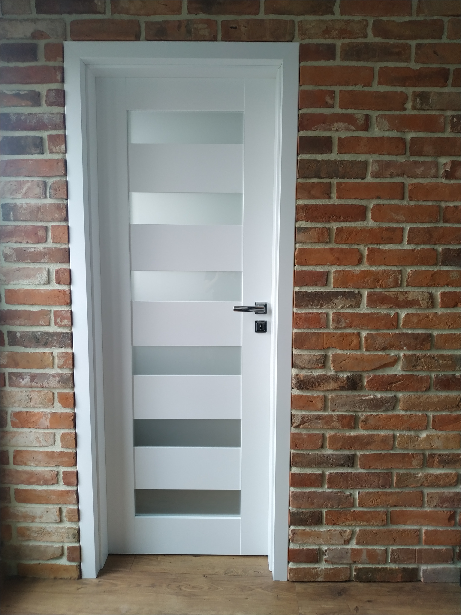 Białe drzwi wewnętrzne z przeszkleniami w wąskich pasach, zamontowane w ścianie z cegły, widok na drewnianą podłogę.