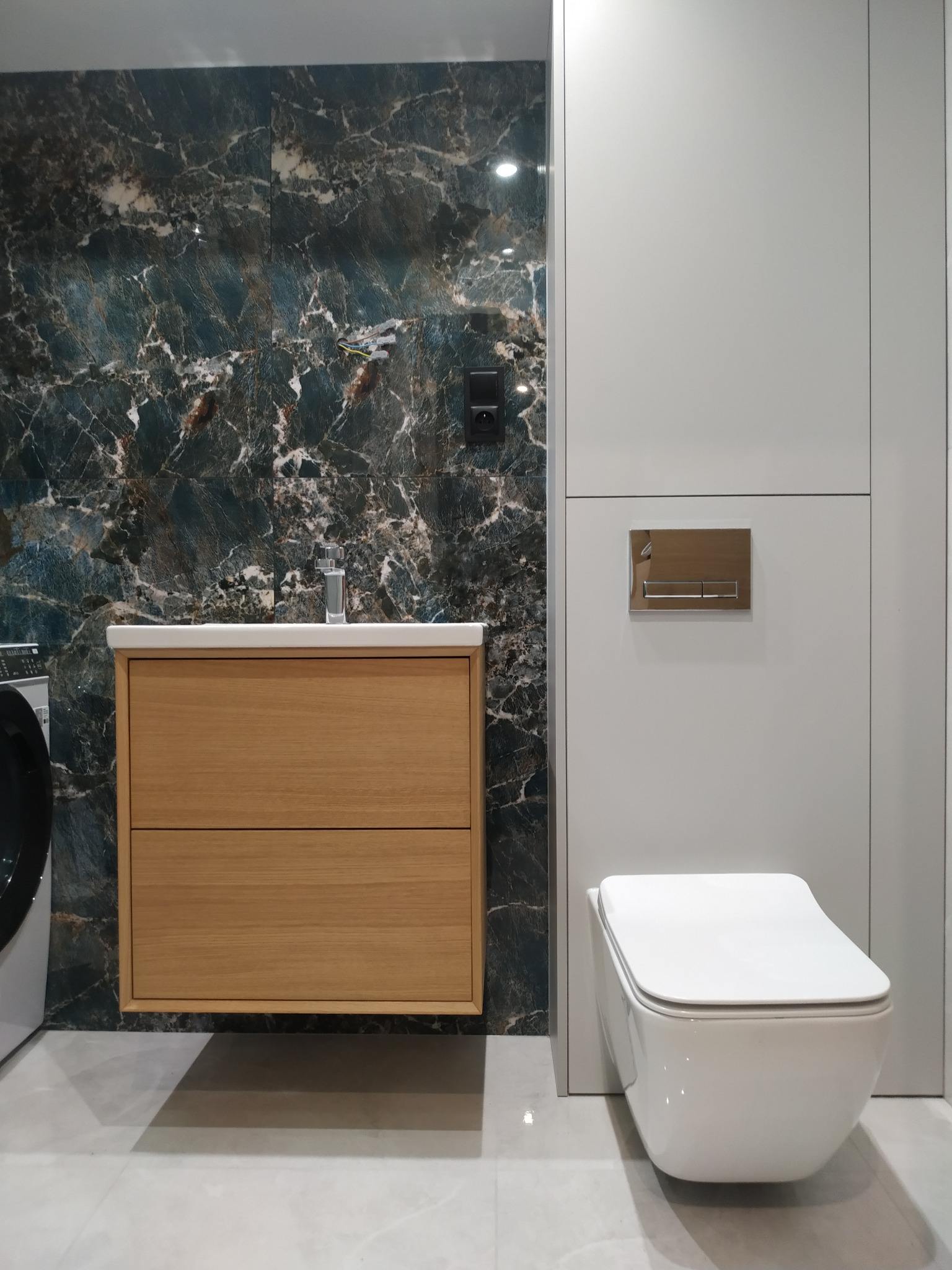 Nowoczesna łazienka z wiszącą toaletą, drewnianą szafką pod umywalką i płytkami imitującymi marmur na ścianie, obok pralka.