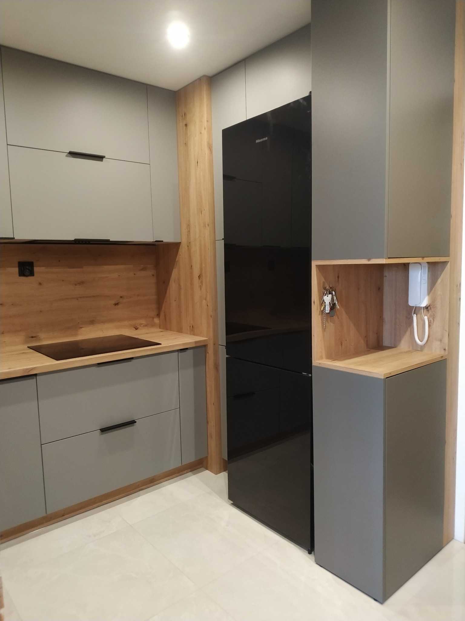 Nowoczesna kuchnia z szarymi szafkami, drewnianymi akcentami i czarną lodówką Hisense. Na blacie płyta indukcyjna, w otwartej półce klucze i domofon.