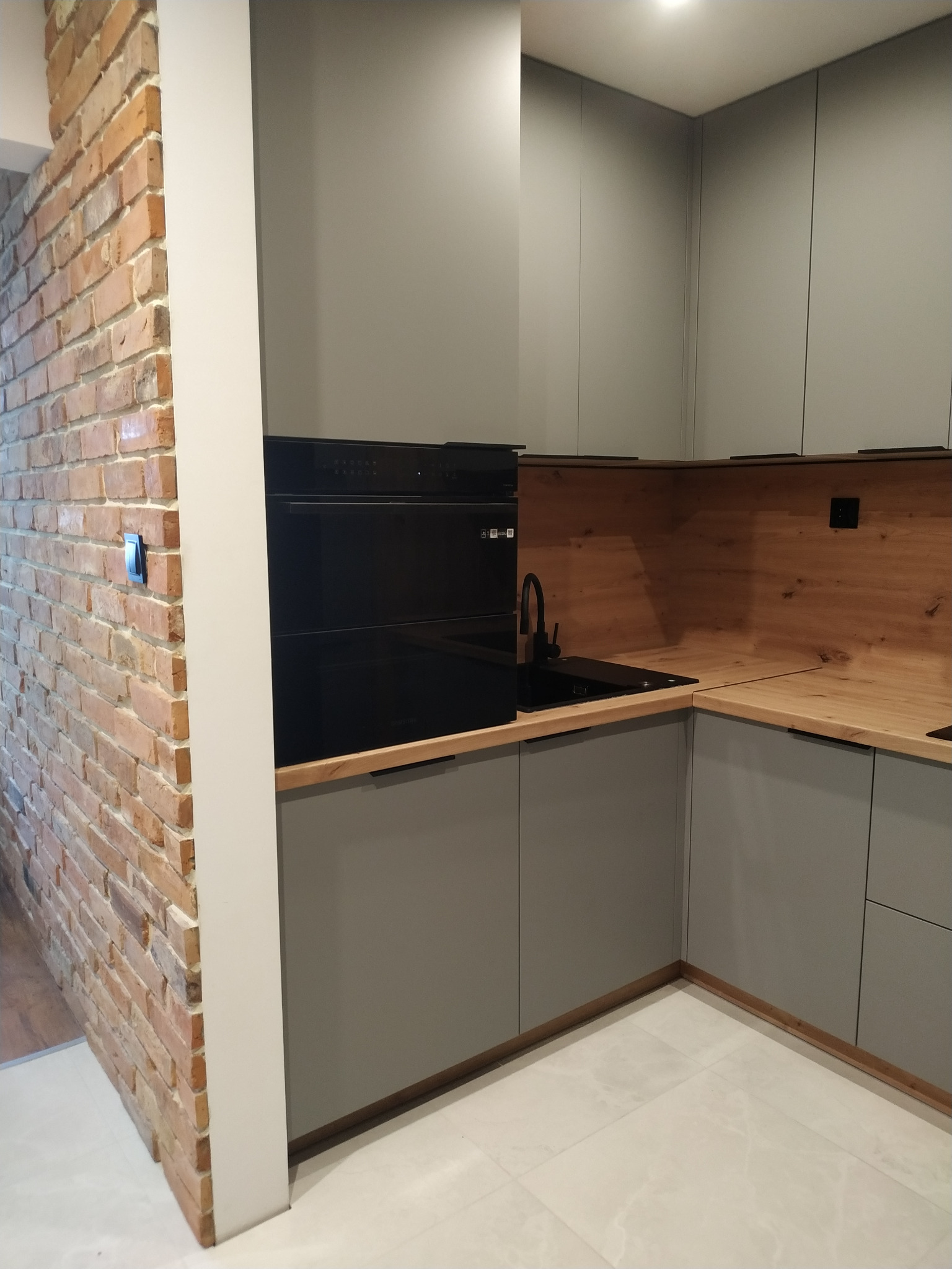 Nowoczesna kuchnia w odcieniach szarości z drewnianym blatem i ceglaną ścianą, czarna armatura i sprzęt AGD, minimalistyczny design, oświetlenie sufitowe.