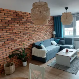 Salon z ceglaną ścianą, szarą sofą narożną, drewnianym stołem kawowym na szarym dywanie, lampami z abażurami z plecionki i rośliną doniczkową. Widoczne także turkusowe zasłony i białe krzesło.