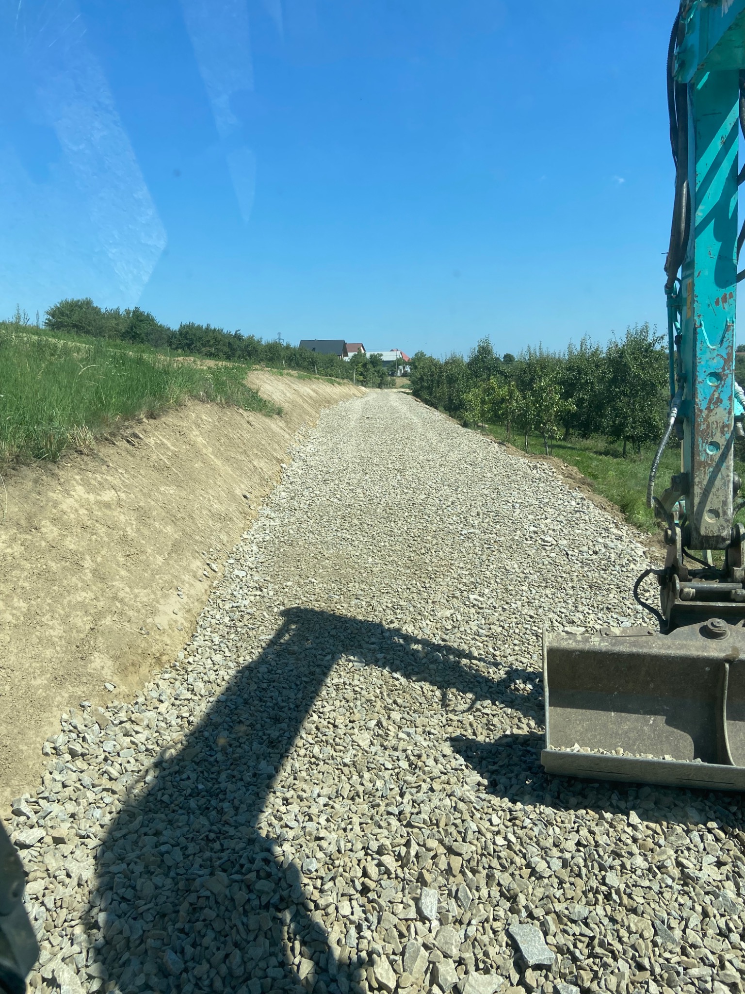 Wyrównana, szutrowa droga w trakcie budowy, widziana z kabiny maszyny budowlanej, z widocznym fragmentem ramienia koparki po prawej stronie. W tle drzewa i zabudowania.