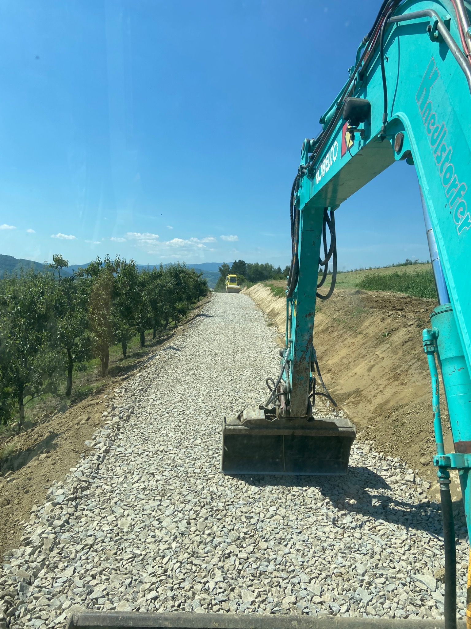 Turkusowy koparko-ładowarka Kobelco na tle drogi z kruszywa, w oddali widoczny walec drogowy i rząd drzew owocowych, błękitne niebo.