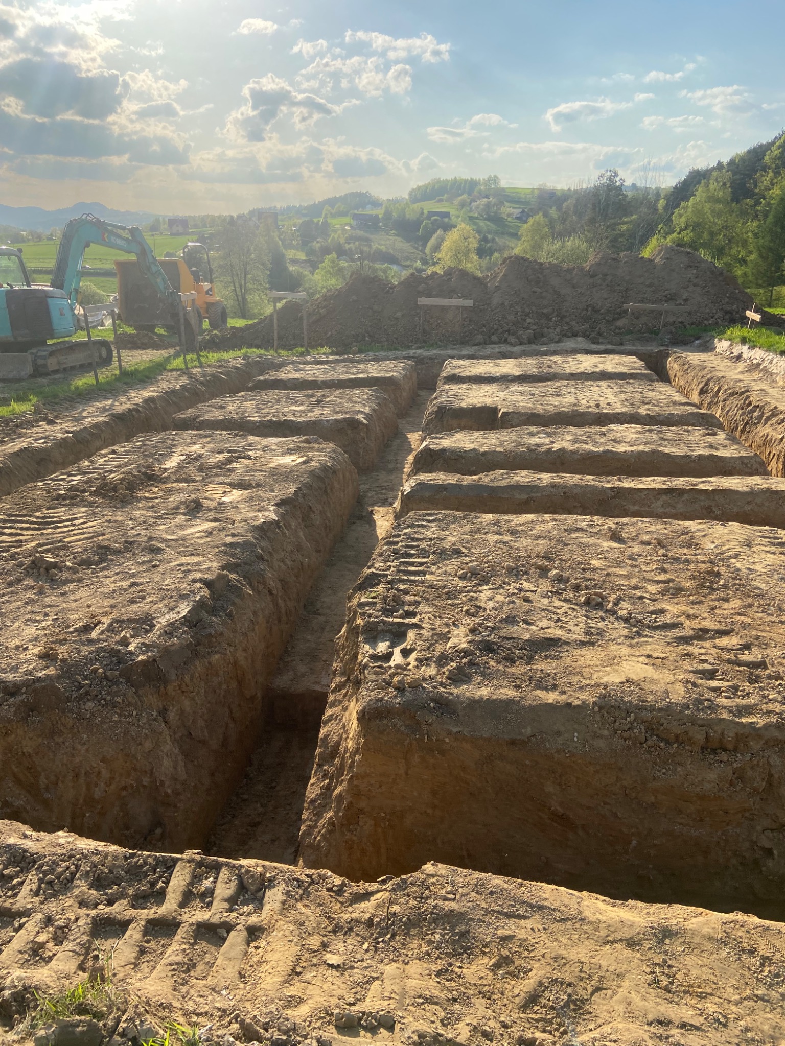 Wykonane wykopy pod fundamenty z widocznymi śladami opon koparki, w tle maszyny budowlane i pagórkowaty krajobraz.