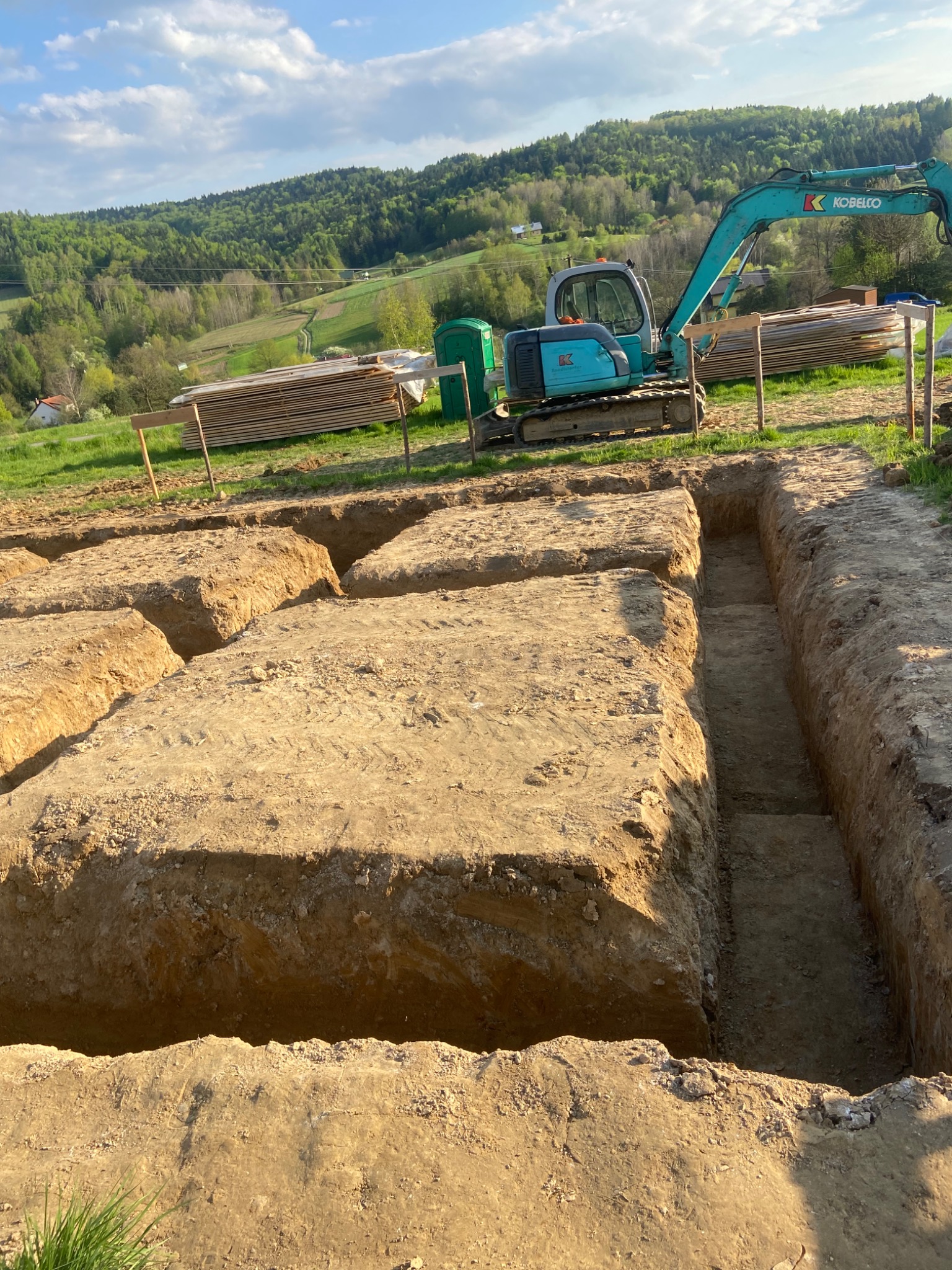 Wykopane rowy pod fundamenty na działce, widoczny błękitny minikoparka Kobelco, stos desek oraz zalesione wzgórza w tle, słoneczny dzień.
