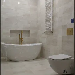 Nowoczesna łazienka w beżowych płytkach, z wolnostojącą wanną owiniętą folią ochronną, złotą baterią, wiszącą toaletą i białym grzejnikiem drabinkowym