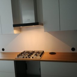 AGNES-STYK ADAM KALMUS - Nowoczesna kuchnia z białymi szafkami i drewnianym blatem, na którym znajduje się płyta gazowa z czterema palnikami oraz okrągła kratka wentylacyjna. Nad płytą widoczny okap marki Samsung.