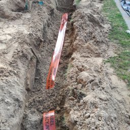 AGNES-STYK ADAM KALMUS - Wykop w ziemi w Szczecinie, zabezpieczony pomarańczową taśmą ostrzegawczą 'Uwaga Kabel Telekomunikacyjny'.