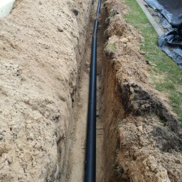 AGNES-STYK ADAM KALMUS - Długi, czarny przewód ułożony w wykopie ziemnym, widok z góry na instalację podziemną.