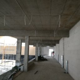 AGNES-STYK ADAM KALMUS - Wnętrze w stanie surowym z widocznymi kablami elektrycznymi zwisającymi z betonowego sufitu, przygotowanymi do dalszej instalacji.