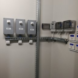 AGNES-STYK ADAM KALMUS - Szara ściana z zamontowanymi szarymi skrzynkami z kluczami, oznaczonymi jako KORT 1, KORT 2 i KORT 3, oraz szeregiem liczników i regulatorów na metalowych szynach.