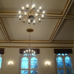 AGNES-STYK ADAM KALMUS - Elegancki żyrandol z wieloma ramionami i żarówkami, zawieszony w przestronnym wnętrzu z wysokim sufitem zdobionym sztukaterią i trzema wysokimi oknami łukowymi z brązowymi roletami rzymskimi.