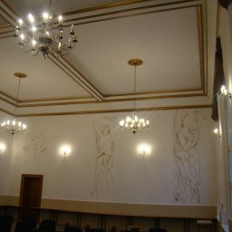 AGNES-STYK ADAM KALMUS - Wnętrze sali z dwoma żyrandolami, kinkietami ściennymi i szkicami postaci na ścianach, złote zdobienia sufitu i ścian, rzędy krzeseł i stołów.