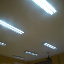 AGNES-STYK ADAM KALMUS - Równomiernie rozmieszczone, podłużne lampy rastrowe na suficie w pomieszczeniu biurowym o żółtych ścianach. Widoczny również projektor przymocowany do sufitu.