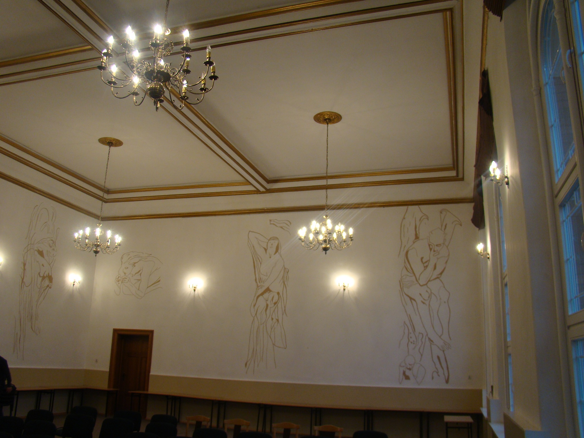 Wnętrze sali z dwoma żyrandolami, kinkietami ściennymi i szkicami postaci na ścianach, złote zdobienia sufitu i ścian, rzędy krzeseł i stołów.