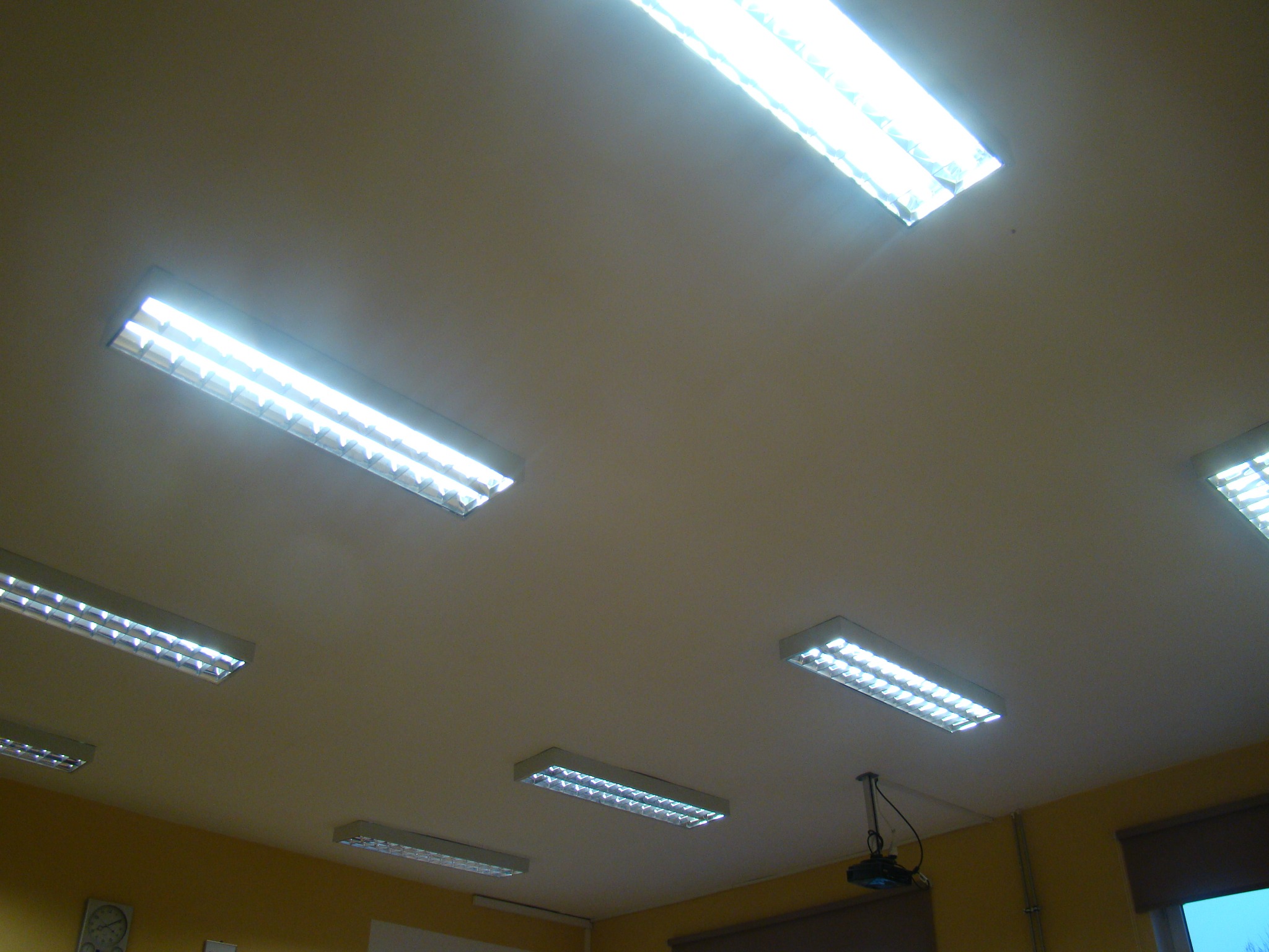 Równomiernie rozmieszczone, podłużne lampy rastrowe na suficie w pomieszczeniu biurowym o żółtych ścianach. Widoczny również projektor przymocowany do sufitu.