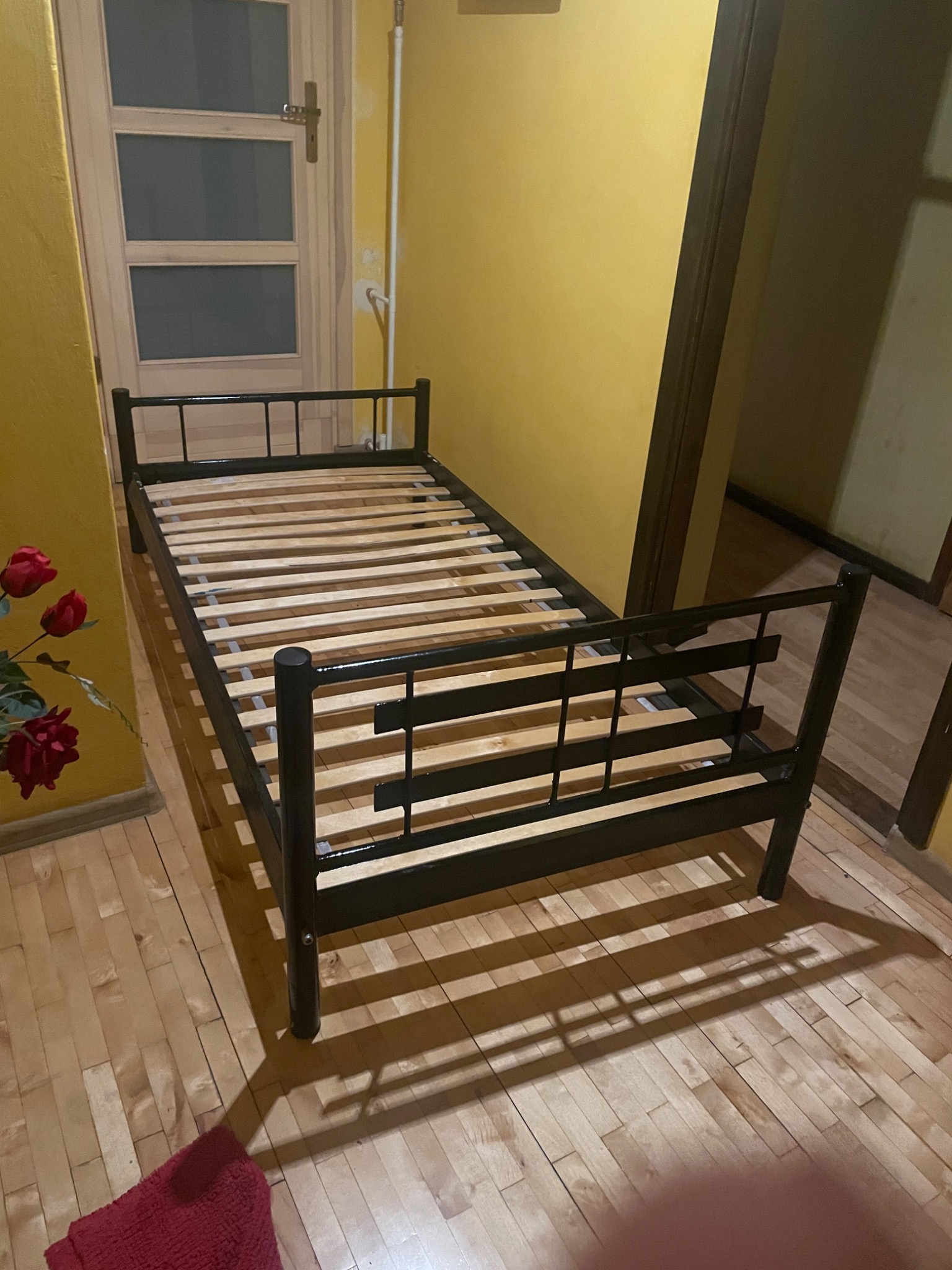 Czarna metalowa rama łóżka z drewnianym stelażem na podłodze z paneli, żółte ściany w tle, obok drzwi i sztuczne czerwone róże w wazonie.