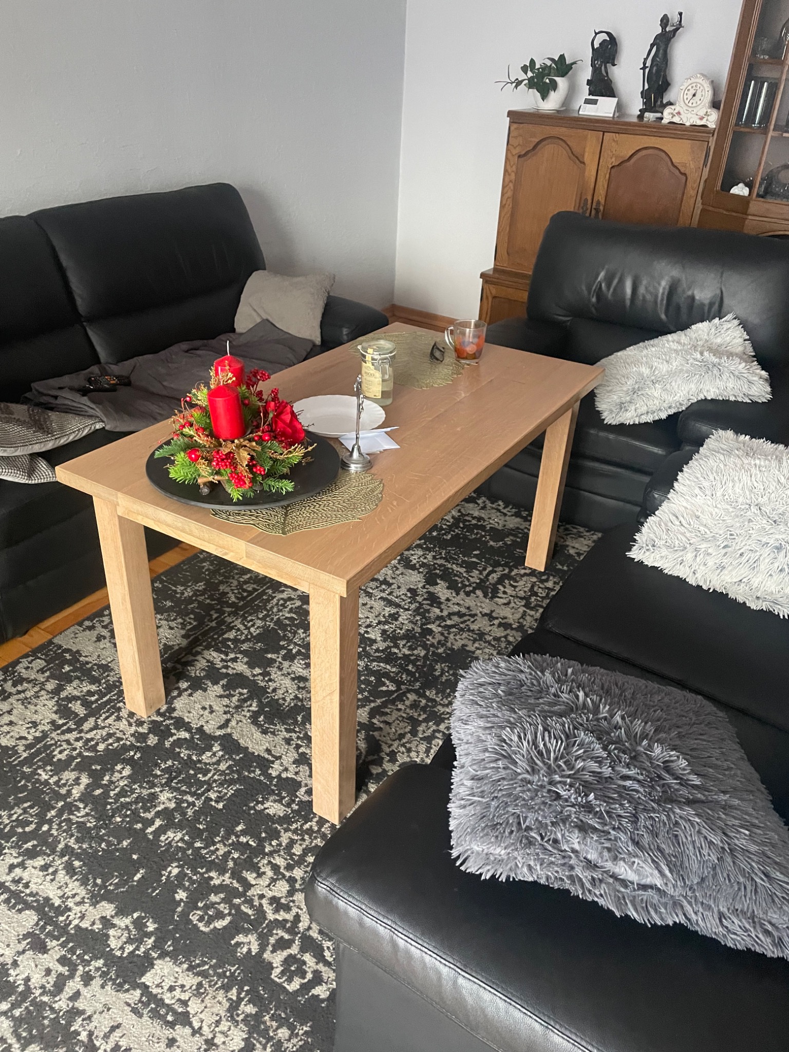 Jasny drewniany stół w salonie z czarną skórzaną kanapą, na stole dekoracja świąteczna z czerwonymi świecami i sztucznymi kwiatami, obok talerz i szklanka.