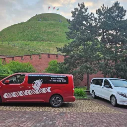 Dwa busy: czerwony z grafiką płomieni i numerem telefonu oraz biały Mercedes Vito, stojące na tle Kopca Kościuszki w Krakowie.