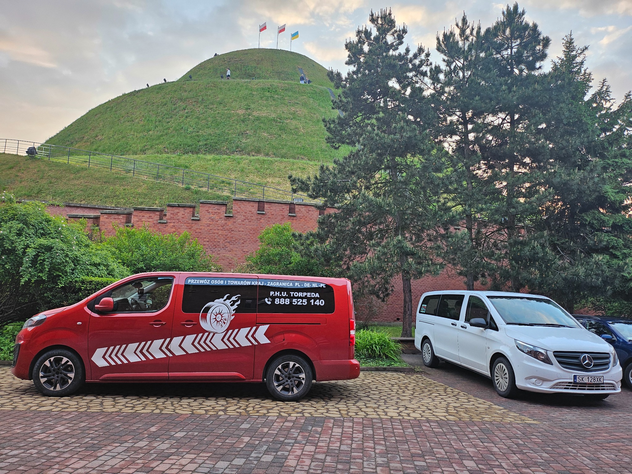 Dwa busy: czerwony z grafiką płomieni i numerem telefonu oraz biały Mercedes Vito, stojące na tle Kopca Kościuszki w Krakowie.