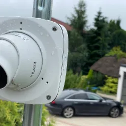 Zewnętrzna kamera monitoringu Kenik, zamontowana na metalowym słupku, z widocznymi kroplami deszczu na obudowie, w tle dom z garażem i zaparkowane samochody.