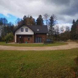 Nowoczesny dom jednorodzinny z cegły klinkierowej z ciemną dachówką, otoczony zielonym trawnikiem i żwirową drogą dojazdową, na tle lasu.